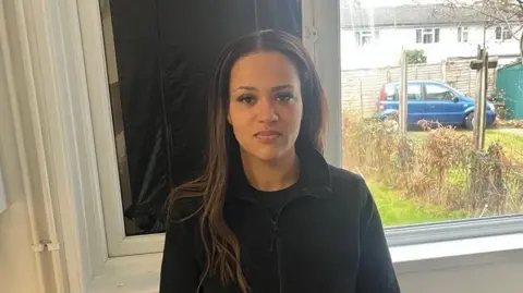 Adrian Harms / BBC A woman wearing a black jumper standing in her home in front of a boarded-up broken window.