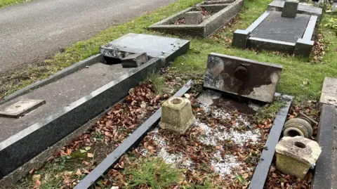 Picture shows several grave headstones uprooted from the ground, knocked over on to the grass. 