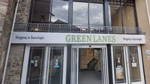 The outside of Green Lanes shopping centre in Barnstaple. It is a brick building with large glass windows and the words 'Green Lanes' above the door. The entrance has large glass panels and on the first floor, there are large glass windows.
