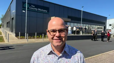 A man in a multi-coloured spotted shirt stands in front of a large black building. "The County Mortuary" is written on the side.
