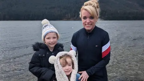 Loretta Davies Loretta Davies has blonde hair (picture back right) and smiles at the camera with her two children stood next to her in front of a lake. Both children wear fluffy hats. 