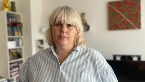 Justine Gordon-Smith stands in a room and has an unsmiling expression as she faces the camera. She has shoulder-length blonde hair with a fringe and wears a shirt with vertical blue and white stripes.