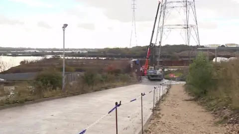 View of the entrance to the Howdon treatment works, with the banks of the River Tyne to the left, a road with a digger at the end of it, next the the lower part of an electricity pylon.