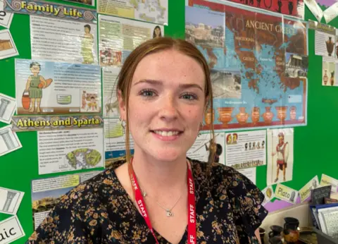 Gemma Dawson/BBC Teaching assistant Lauren Brown. She is wearing a flowery dress and has tied back auburn hair. She is standing in a classroom and has a red staff lanyard around her neck.