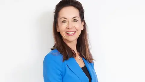 Lizzy Bowen with shoulder-length brown hair, a blue blazer and black top standing in front of a white background smiles at the camera. 