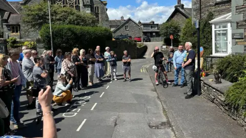 Laszlo Papp Over a dozen people standing on a road on a sunny day crowding around and taking a photo of several men next to a cat statue