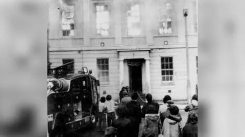 Newry Reporter A black and white newspaper photo of the former Newry Savings Bank on fire after a bomb attack in 1972. A fire engine is parked outside as a crowd of people look up at flames coming out of the top-floor windows. 