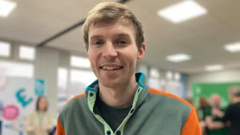 David Barclay wearing a green fleece pullover with orange and blue detailing. He has short blonde hair and blue eyes, and is smiling at the camera. Behind him there are tables and banners set up with information on how to reduce energy costs and where to find support. 