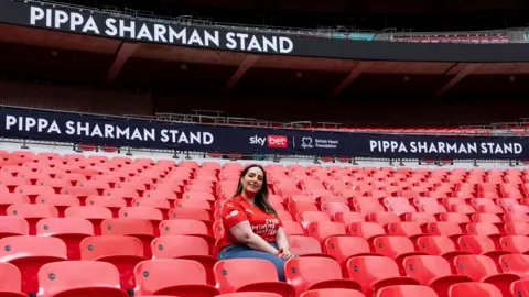 BHF A picture of Pippa Sharman, wearing a red Every Minute Matters T-shirt, sat in Wembley stadium with "Pippa Sharman Stand" behind her on screens running behind her.