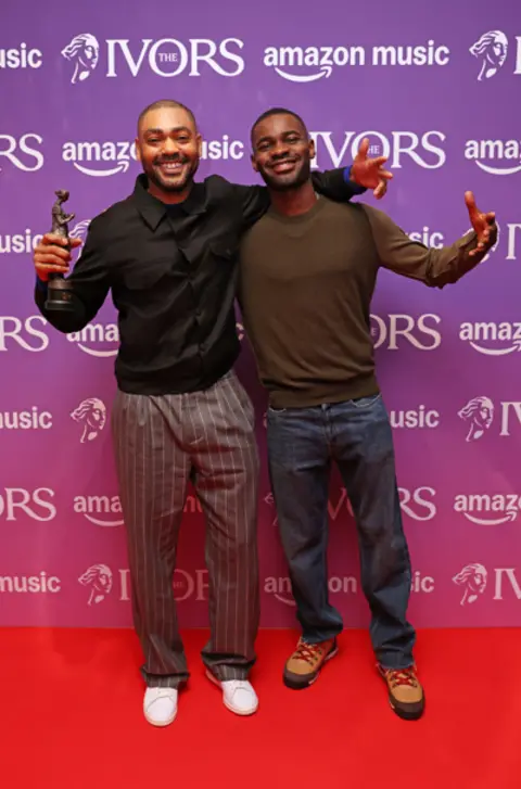 Getty Images Kano and Dave smile and stand on the Ivors red carpet. Kano holds his Ivor Novello Award.