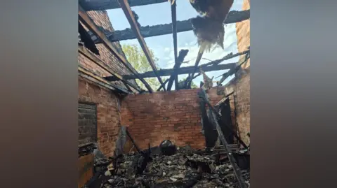 Jade Fenton A damaged house including a missing roof and lots of damaged, burnt items on the floor and an exposed brick wall. 
