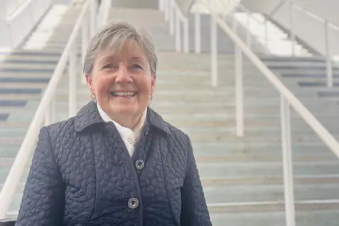 Joyce Keenan smiling at the camera at bottom of steps, she is wearing a blue padded jacket.