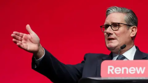 Reuters Sir Keir Starmer is speaking at a lectern which has the word "Renew" written on a sign attached to it. Sir Keir is wearing a dark coloured suit, a white shirt, a tie and glasses. His right arm is outstretched as he speaks in front of a red background.