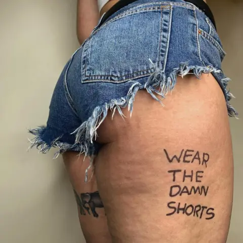 Georgina Cox Instagram post by Georgina Cox, showing her body from the waist to mid thigh. She's wearing a pair of very short cut-off jeans shorts, with a frayed hem. A little of her bottom is showing and on her average-sized and strong-looking upper thigh are the words "Wear the damn shorts".