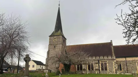 Babergh District Council St Andrew’s Church in Great Cornard, gravestones and trees can be seen in the foreground, the church is in the middle of the image with a brown slated roof and tall dark spire. The walls have light stonework.