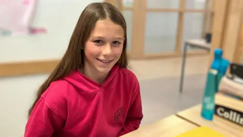 Etta sitting at a classroom desk wearing a deep pink hoodie.