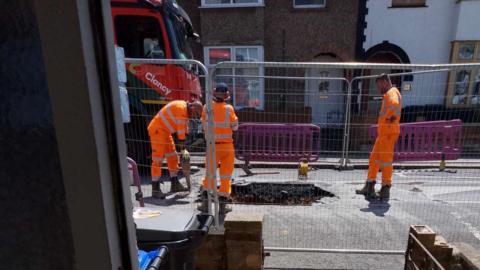 Three workers in orange overalls look at a large hole in the street. The hole is surrounded by barriers.