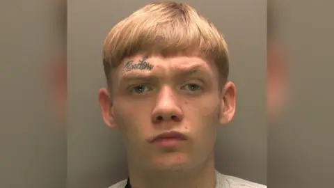 A police photo of a man with short blond hair looking seriously into the camera with a frown. He has a black tattoo that reads "freedom" above his eyebrow.