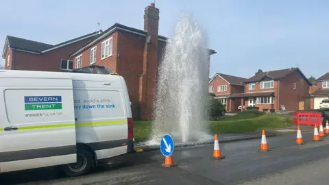 A different angle showing water shooting into the sky with a Severn Trent van parked nearby and cones marking out the affected area. A row of houses are pictured in the background.