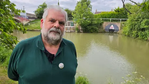 Graham Puddephatt stands on the banks of the River Kennet. He has white hair and a beard and wears a green polo shirt.