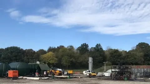 The drilling site at Horsehill in Surrey, viewed from a distance, with two cylindrical green tankers, equipment and vehicles, and a line of trees in the background.