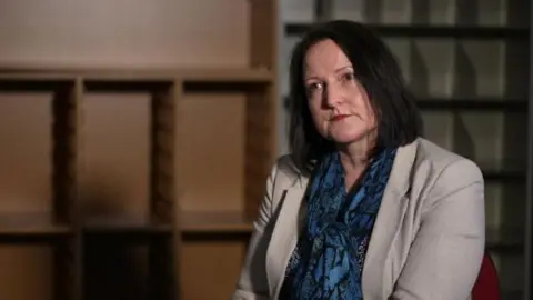 Alison Hernandez wearing a blue, snake skin top, with a cream. She has shoulder length black hair. Ms Hernandez is sat on a chair and it looking to the left of the camera. 