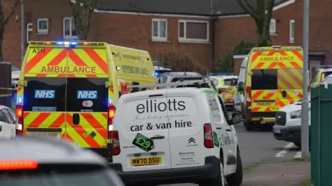 Two ambulances can be seen on a small road with parked cars, surrounded by houses