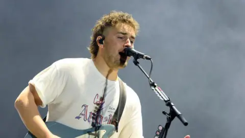 Getty Images A photo of Sam Fender holding a blue electric guitar and singing into a microphone, wearing a white tshirt 