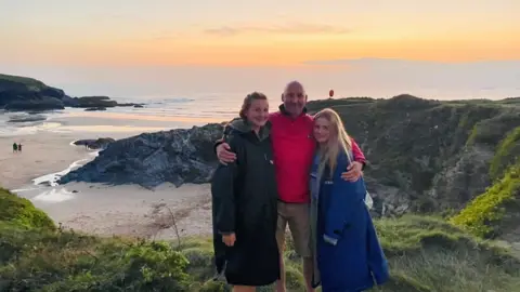 Shaun Lyle A man and two girls stand on the side of a cliff, which overlooks the beach and the sea.