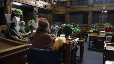 A group of people sat in a council room dressed in various nature inspired costumes, including a tree