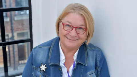 Rosemary Shrager stood in front of a white wall and black panelled window frame. She is wearing a blue denim jacket, white shirt and red-framed glasses.