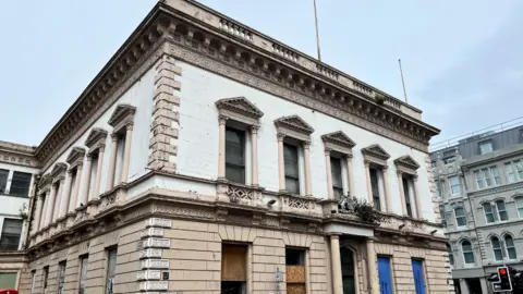 Exterior of large stone, derelict building. It has numerous ornate windows, some of which are boarded up with wood and blue signs 