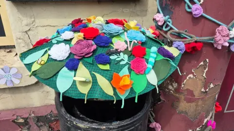 Wem Town Council A black public waste bin which has a knitted and crocheted display on top. There are green leaves and colourful flowers, as well as a black spider, bugs, bees and butterflies