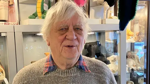 Bernie Jones, a man with white hair and a grey jumper standing in front of cabinets of soft toys