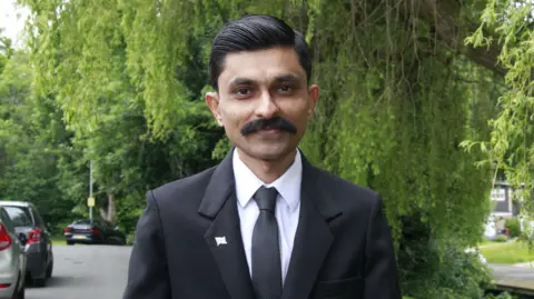 Submitted Mr Pipaliya smiles for the camera while standing in a tree-lined street. He has a moustache and short, sleeked black hair. He is wearing a black suit, tie and white shirt. 