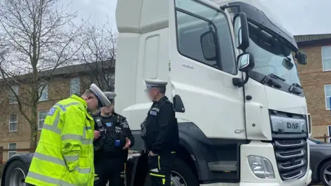 Essex Police Three uniformed police officers stand next to the cab of a white lorry. The lorry is parked outside a block of flats.