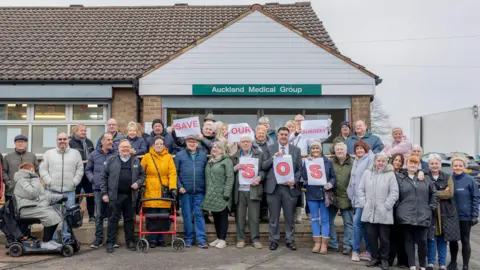 Sarah Caldecott A group of people are standing outside the GP surgery holding placards.