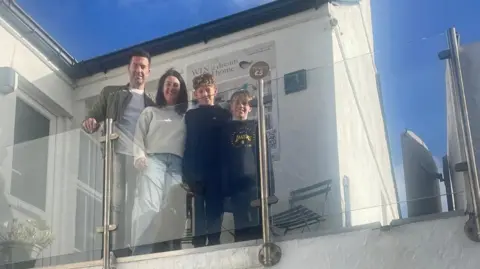 Jennie Bailey A man, woman and two boys stood on a balcony with a white house behind them. They are all smiling and looking at the cmaera.