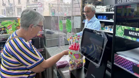 Kate Bradbrook/BBC A woman stands at a shop counter reaching past a jar of lollipops, with a clear protective screen between the counter and the cashier