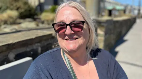 Trish Roberts has fair blonde hair tied up with lose pieces falling by her face. She is wearing large sunglasses, and a dark blue cardigan, with a strap of a bag visible on the left over her shoulder. She is smiling sitting in front of a long stone wall with a pavement to the right behind her, on a sunny day.
