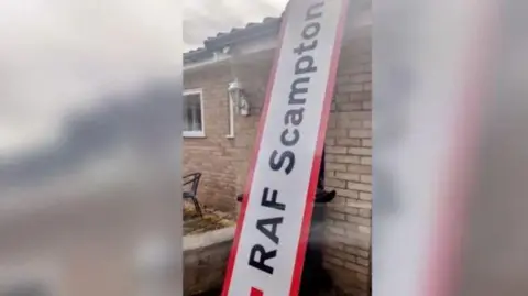 Steve Jollands A road sign with ‘RAF Scampton’ written in black letters can be seen leant against a brick bungalow, it is oversized against the house. 