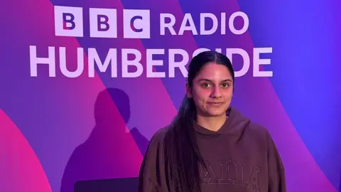 Charan Kaur Dhesi looking at the camera. She has long black hair tied at the back and is wearing a dark hooded top and is standing in front of a multicoloured BBC Radio Humberside banner