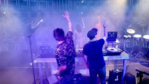 Colin Moody Two male DJs standing on stage at Bristol Harbour Festival with their arms in the air, hyping up the crowd below. The amphitheatre is full of a large crowd of people. The sky is dark and the image has an overall blue-purple hue from the stage lights. 