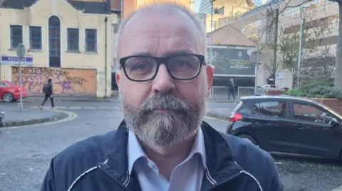 BBC A man with short balding grey hair and a grey beard, looking at the camera. He is wearing glasses, a navy jacket and a blue shirt.
