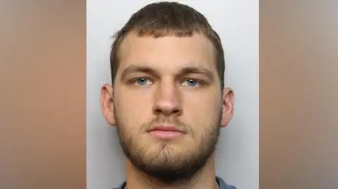 Northamptonshire Police The custody photo of a man who has brown hair and a beard. He is looking directly at the camera and is wearing a blue shirt.