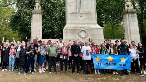 About 80 walkers are pictured standing in front of Southampton Cenotaph in the city centre. They are wearing comfortable hiking clothes and smiling and waving to the camera. A blue banner inscribed with the names CJ, Mario and Vlad is being held by their families.