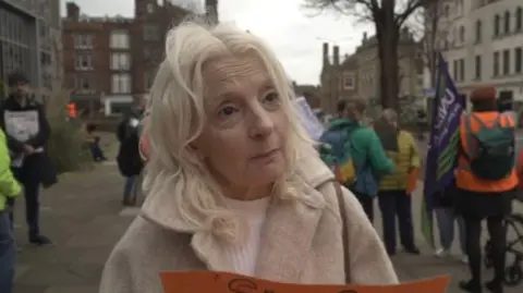 LDRS/Oliver Leader de Saxe A woman with light blonde hair, wearing a beige-coloured coat and holding an orange poster. She is standing near a group of protesters outside a local council building.