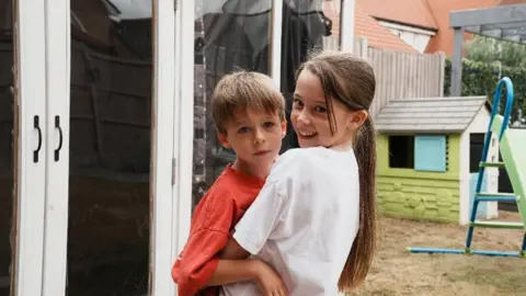 John Fairhall/BBC Freddie being embraced by his sister while she turns to smile to the camera. There is a Wendy house and a slide in the background.