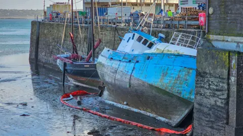 The Karina Olsen leaning against the harbour wall in Penzance. It has a blue hull around which is a red oil boom. The tide is out and the revealed sea bed looks muddy. There is a smaller boat moored upright to the left of the former trawler. There are vans, wooden pallets and metal containers lining the top of the harbour wall along with a couple of people. 