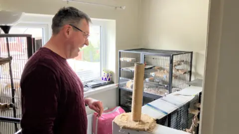 A man in a burgundy jumper and glasses, smiling as he looks into a room containing a number of large metal cages with shelves in, as well as a metal framed pen on the floor